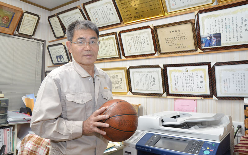表彰状や感謝状をバックにバスケットボールを手にする時吉コーチ